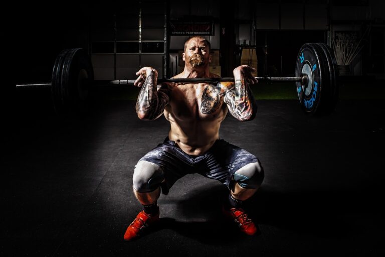 On voit un homme musclé en train de soulevé une barre sur ses clavicules en faisant un squat dans un environnement sombre